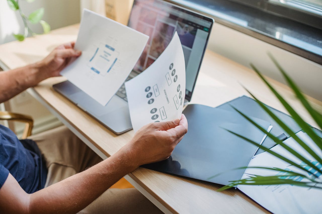 digital Crop anonymous male designer in casual wear sitting at table with opened laptop and folders and reading papers